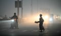 A woman walks across a street engulfed by smog as headlights from a nearby motorbike and other vehicles behind shine into the gloom