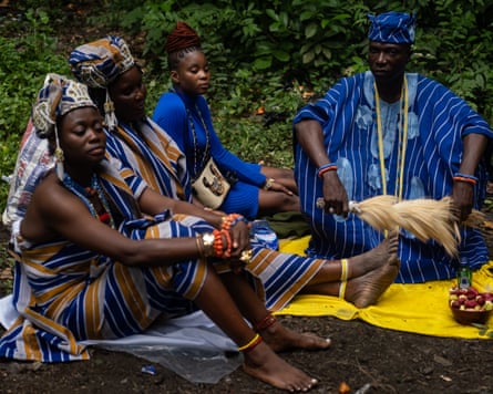 Three African women and a man in traditional dress sitting on cloths with kola nuts and a small bottle in front of them
