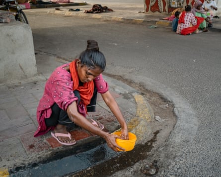 A woman crouches on a kerb, holding a small yellow plastic bowl. Water can be seen in the gutter