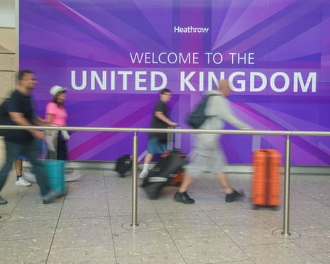 Travellers with luggage walk past a purple wall reading 'Welcome to the United Kingdom' at Heathrow airport