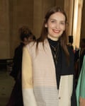 Roksanda Ilinčić, photographed at London fashion week, February 2022: she is wearing a black high-necked top with three gold necklaces, the lowest one a cross, under a collarless oversized jacket with panels in shades of pale gold, cream and grey with narrow stripes and a shoulder in black; it is fastened with a single button at waist level. She has straight, brown hair worn loose and is wearing bright red lipstick and smiling at the camera.