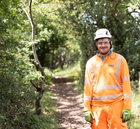 A man in an orange HS2 work uniform and white hard hat stands for a picture on a wooded track.