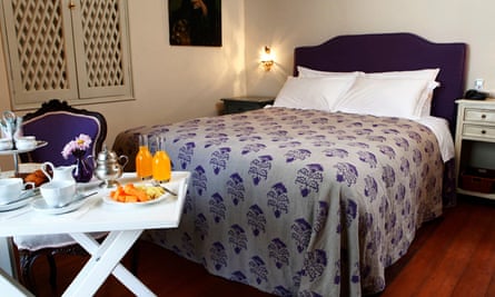A bedroom at Quinta Miraflores B&B in Lima, Peru; a purple bedspread adorns a bed in a predominantly white room. A breakfast tray with orange juice and fruit is in front of the bed.