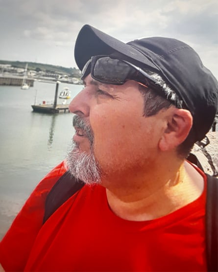Claudio Aquilino wears a black peaked cap and sunglasses pushed up on his forehead. He has a greying goatee beard and round face. He is standing at a waterfront with a harbour and small boats behind him.