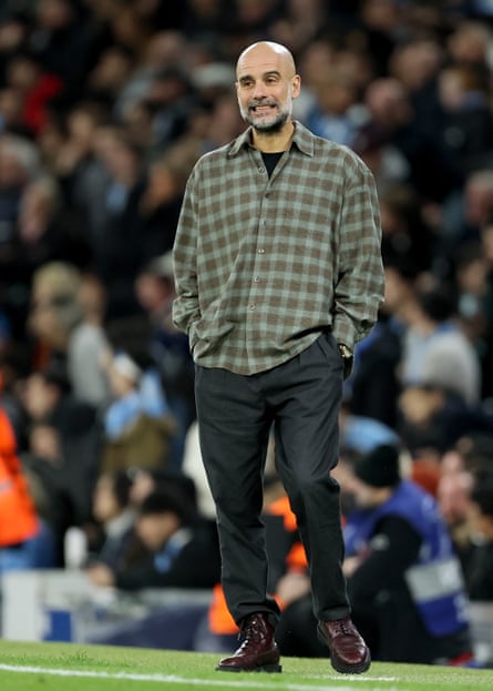Pep Guardiola wearing a checked shirt and black trousers on the sideline of the pitch