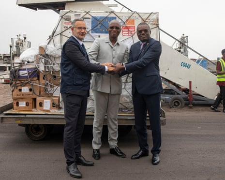 Three men shake hands in front of a loading pallet piled with boxes.