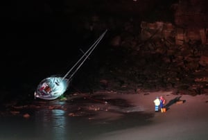 A sailing boat at an angle in the bay. Two emergency services workers are standing on the sand looking at the boat
