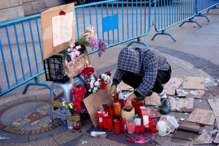 Person with hood of jacket over head crouches down at makeshift shrine with candles and flowers next to blue crowd barriers