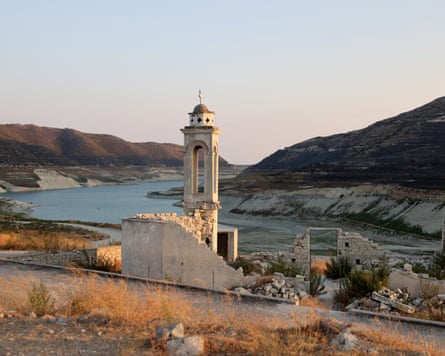 The bell tower of a white church with tumbledown walls stands by a road with a shallow stretch of water between hills behind. It appears to be on dry land with grasses, reeds and small bushes around it.