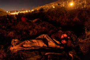A woman and her young daughter wrapped in a blanket bathed in an orange light. Lights at a border fence are behind