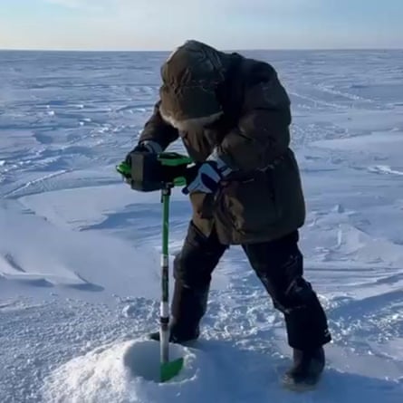 A person wearing heavy outdoor gear stand in a flat snowy landscape bearing down on a metal pole the end of which is buried in the snow in front of them.