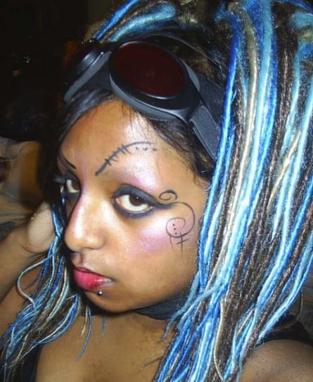 A close up sideways view of a young South Asian woman with blue streaky hair, and a pair of goggles on her head, wearing brightly coloured makeup and with markings on her face
