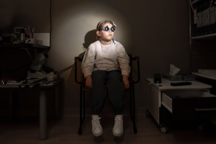 A young girl in a white top and white trainers sits in darkness at an oculist’s, with her face illuminated. She is wearing surgical glasses for tests that look like goggles.