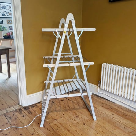 A folded three-tier heated clothes airer stands on a wooden floor near a radiator