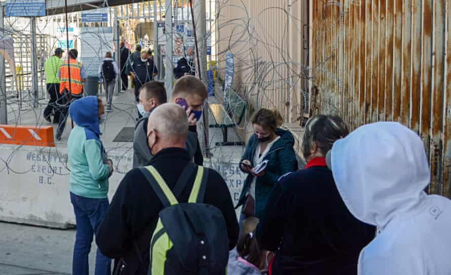 A group of people carrying backpacks and wearing masks wait at a barbed-wire barricade in front of a border-crossing gate.