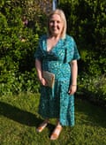 Rebecca wears a green patterned dress and orange sandals, and holds a cream clutch bag, as she stands in a garden smiling. She has shoulder-length straight blond hair.