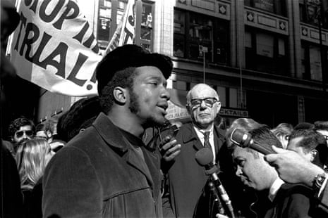 Black and white photograph showing a black man talking to white men holding microphones, while behind him a older white man looks on. Others hold banners in the background.