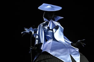 A woman in an elaborate silver coat and wide-brimmed hat carrying a silver flower