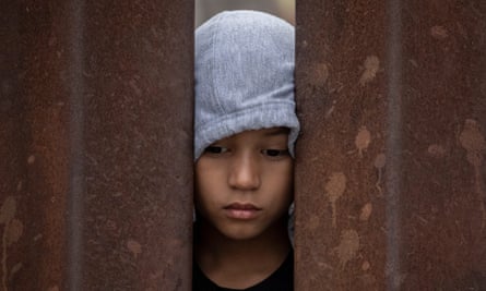 Young boy with brown skin and gray hoodie presses his face to rusty bars and looks downward.