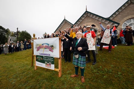 People stand outside the stone village hall and raise tiny shot glasses of whisky. Some are wearing aprons and others kilts.