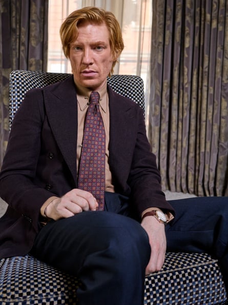 A man sitting on a chair in a blue suit, wearing a red shirt and dark red patterned tie