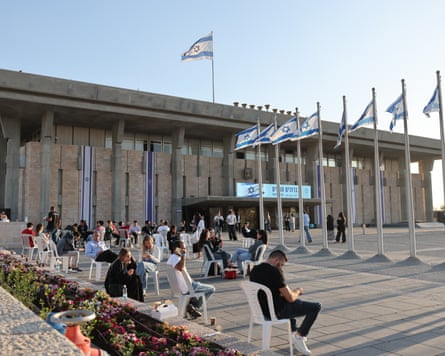 People sit on plastic chairs outside Israel's Knesset parliament building, with Israeli flags flying nearby