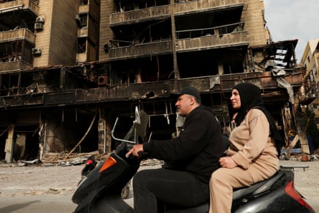 A man and woman on a motorbike ride past a building destroyed by an airstrike.