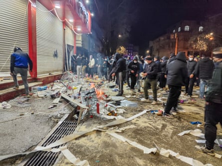 People wearing dark clothing, many with hoods up, gather in front of a closed shop. The ground is littered with rubbish including shopping trolleys