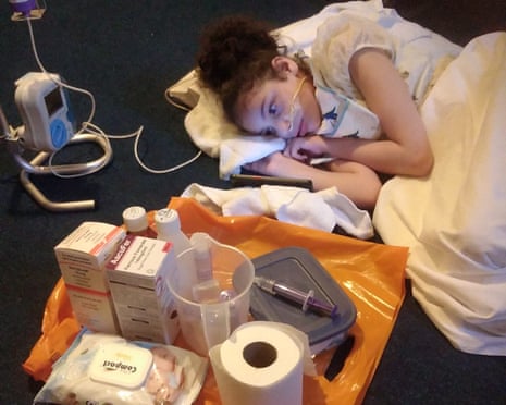 A child lies under a blanket on the floor, with medical supplies and equipment on a tray nearby