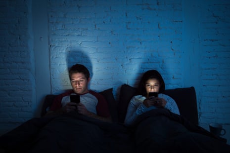 A young couple at home in bed late at night using mobile phones instead of reading