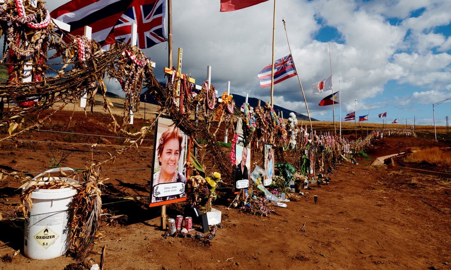 Along a wire fence are fastened a long row of white crosses, flags and images, with one woman’s smiling face most visible.