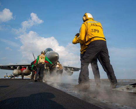 A crew member in yellow vest signals on an aircraft carrier deck as a fighter jet prepares to launch