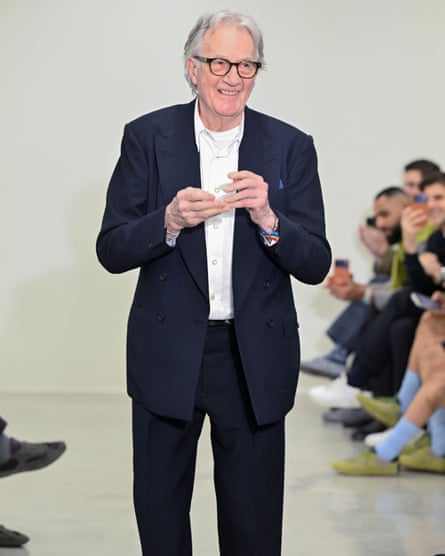 Paul Smith smiling in an open-neck white shirt and dark suit, standing between seated rows of show viewers
