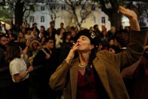 A woman in a jacket smoking a cigarette and dancing at night, as crowds of people stand in the background