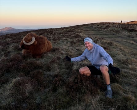Nick Harris-Fry crouches down next to a highland cow on a remote hill in winter