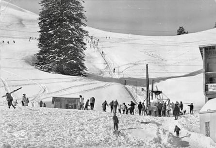 A black and white picture showing skiers gathered at the bottom of a piste thickly covered in snow.