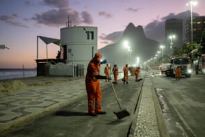 Workers in orange uniform sweep paths and a cycle land beside the beach. A twin-peaked hill is in the background