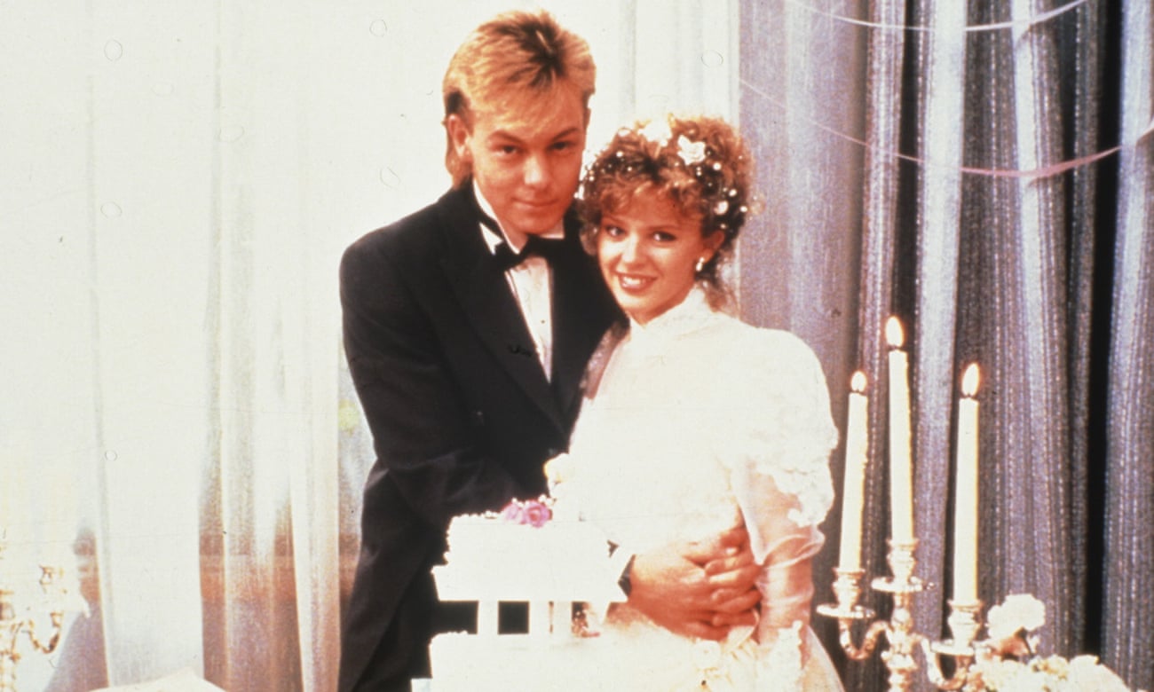 Video still of Scott and Charlene ion their wedding attire embracing, with a wedding cake int he foreground