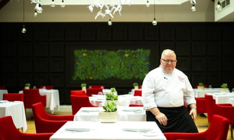 Paul Askew in chef's whites sitting to the right of the photograph by one of the geometrically arranged tables with white tablecloths and red chairs in his dining room