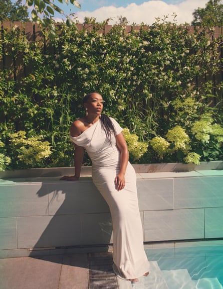 Naomi Ackie stands in the shallows of a pool and wears a white dress and gold pearl drop earrings