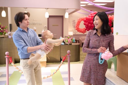 A man in blue shirt and trousers holding a baby doll, with the baby reaching out to a woman wearing a red dress and holding a giant safety pin