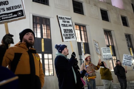 People wearing warm winter coats and hats hold up placards protesting against Elon Musk.