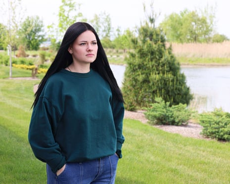 A woman stands on a lawn beside a lake with her hands in her pockes.