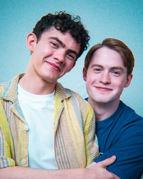 Two young men pose together smiling against a blue backdrop, one in a striped shirt, one in blue.