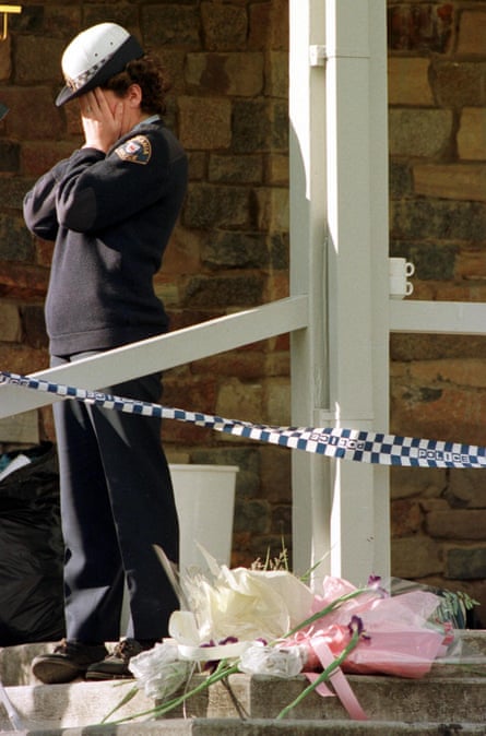 A police woman holds her hands to her face at the top of a set of steps, with flowers resting near her feet and police tape draped from the railing