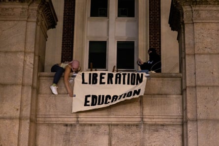Two people unfurl sign that says Liberation Education outside window of stone building at night.