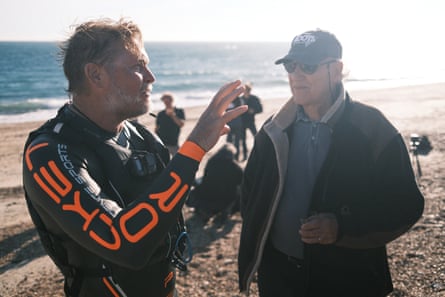 The men talk on a sunny beach, with Bushby in a wetsuit