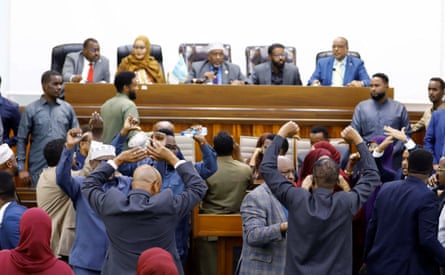 African men in suits stand while making a thumbs-down gesture of protest towards people seated at the front of a parliamentary chamber