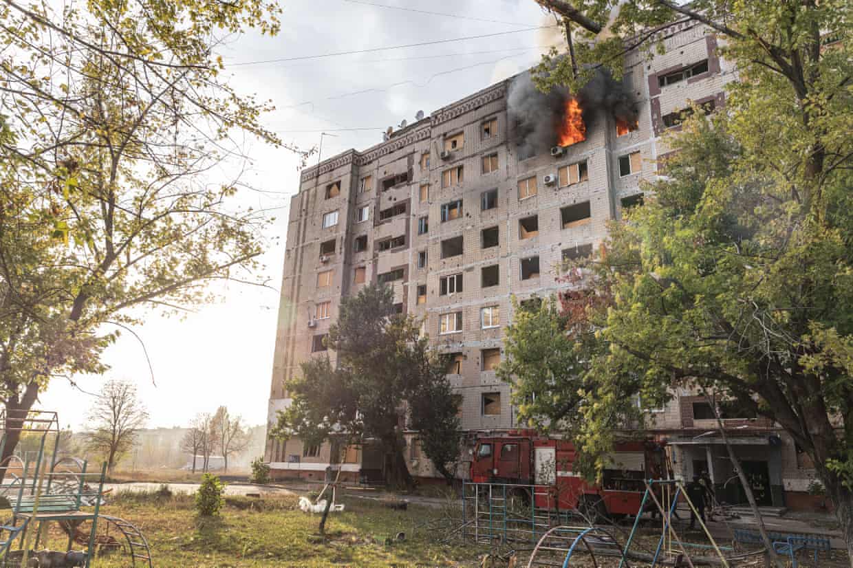 Firefighters attempt to put out a fire in a residential building. Flames and smoke and billowing from windows on an upper floor of a large concrete block.