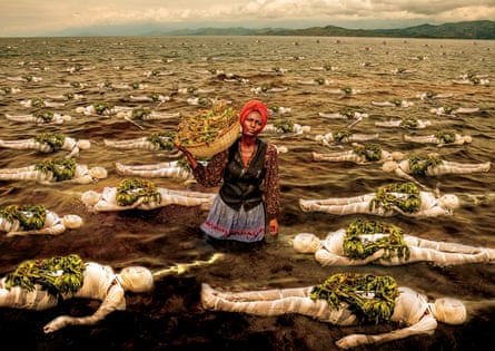 A woman with a basket of leaves on her shoulder stands in a lake surrounded by floating women’s bodies wrapped in bandages with wreaths on top of them made from the same leaves, possibly tobacco